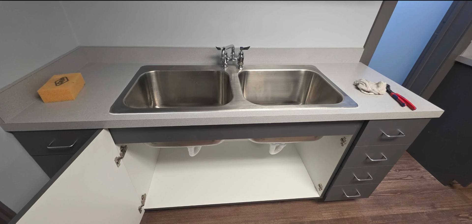 New stainless double-bowl sink and faucet installed in a break room counter with the drain lines finished underneath